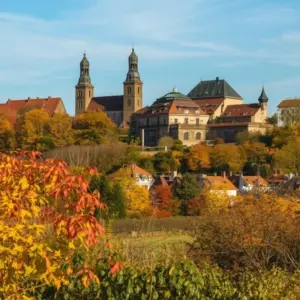 Scenic view of a historic European town featuring a hilltop abbey with twin towers, surrounded by vibrant red and orange autumn foliage.