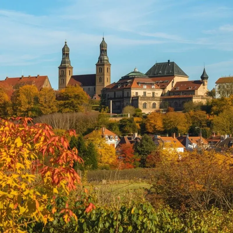 Scenic view of a historic European town featuring a hilltop abbey with twin towers, surrounded by vibrant red and orange autumn foliage.