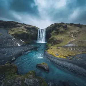 A majestic waterfall cascading over dark cliffs into a winding blue river in Iceland, surrounded by mossy green hills and rocky terrain.