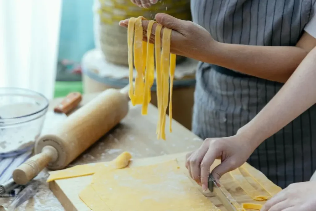 Close-up of hands cutting and flouring long strips of fresh handmade pasta dough on a wooden cutting board.