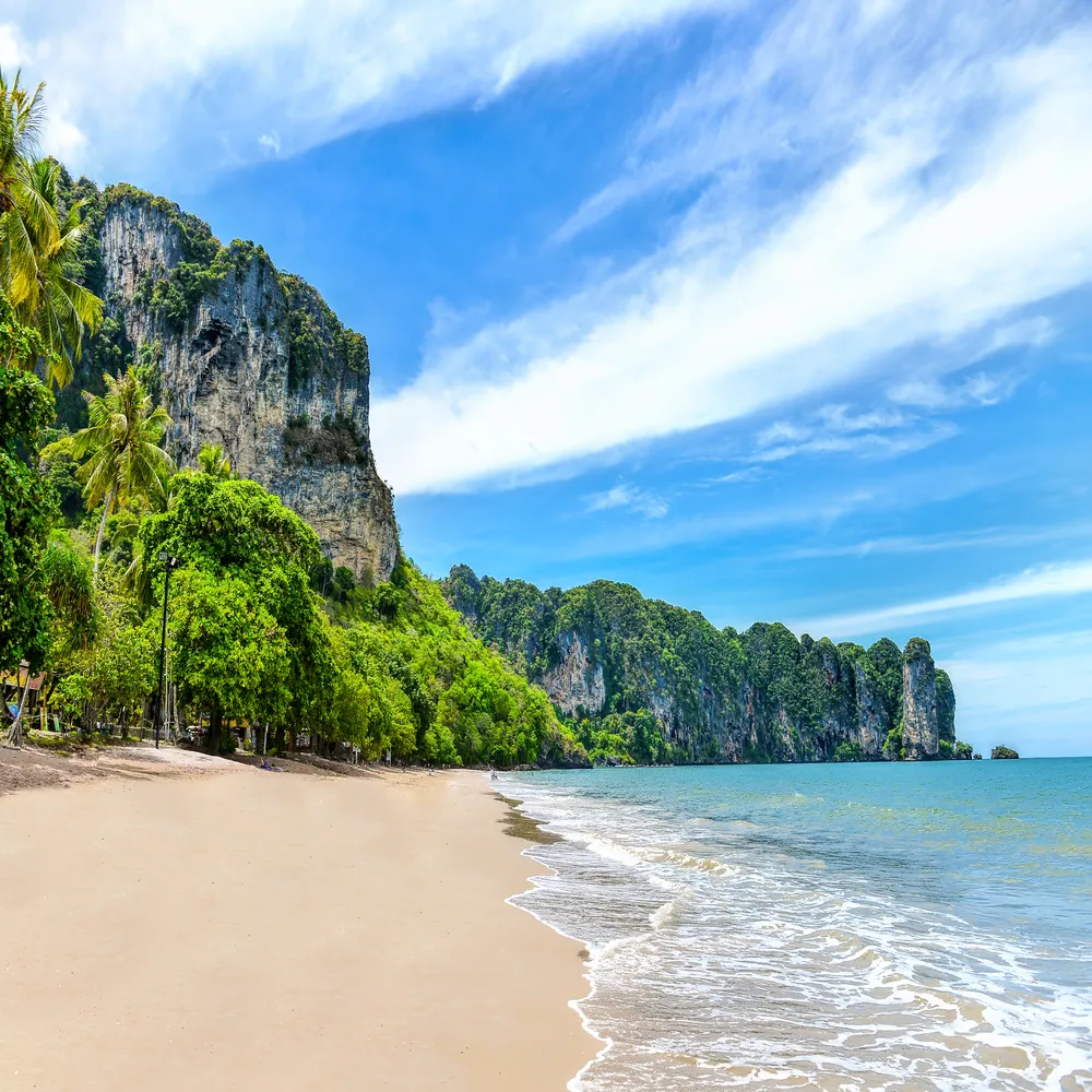 Sunny Ao Nang beach in Thailand featuring white sand, clear turquoise ocean water, and towering limestone cliffs covered in lush tropical greenery.