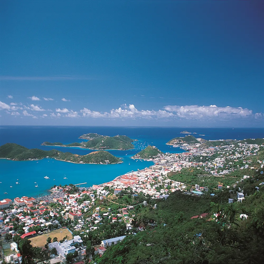 Panoramic aerial view of a Caribbean bay and coastal town from a hilltop, showing bright turquoise waters, distant green islands, moored boats, and colorful island architecture.