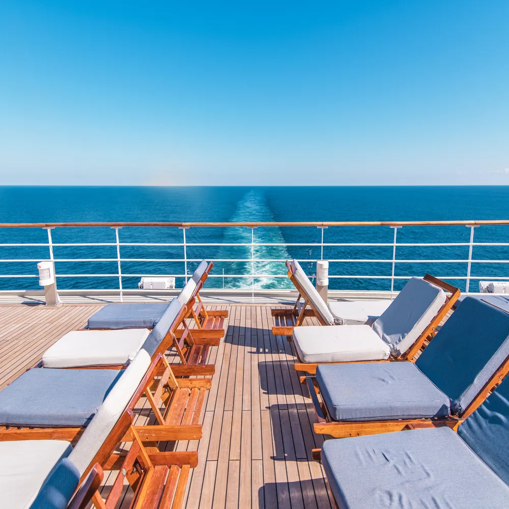 Relaxing wooden lounge chairs lined up on the open aft deck of a cruise ship, overlooking the ship's white water wake trailing into the vast blue ocean.