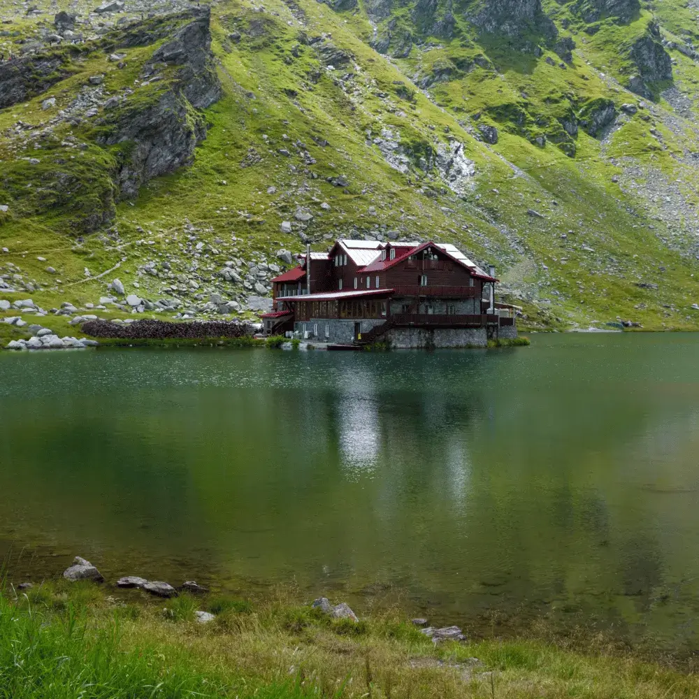 A large wooden chalet resting on the edge of the calm, dark waters of Balea Lake in the mountains of Romania.