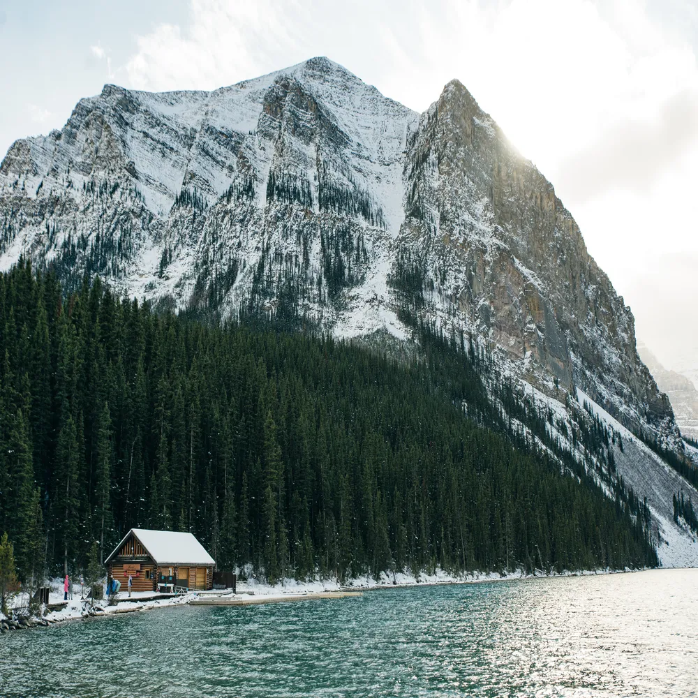 Majestic view of Lake Louise in Banff National Park, featuring a classic wooden boathouse on the turquoise water's edge backed by dense pine forests and a massive snow-capped mountain peak.