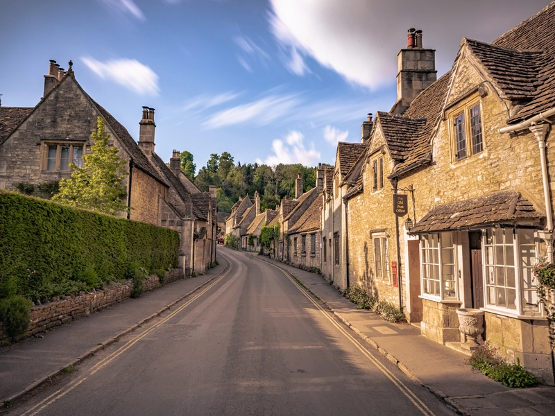A quiet street lined with historic stone cottages and lush greenery in Castle Combe, the Cotswolds, England.