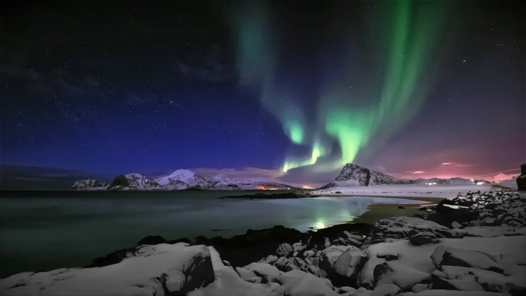 Vibrant green Northern Lights glowing over a snow-covered beach and rugged mountains at night.