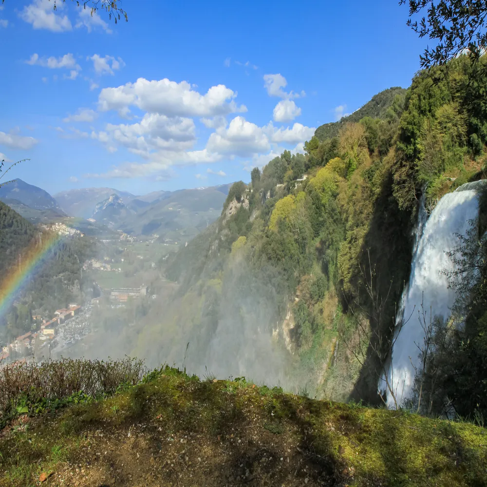 Spectacular, powerful waterfall cascading down a steep, lush green mountainside into a valley, with a bright rainbow forming in the mist below.