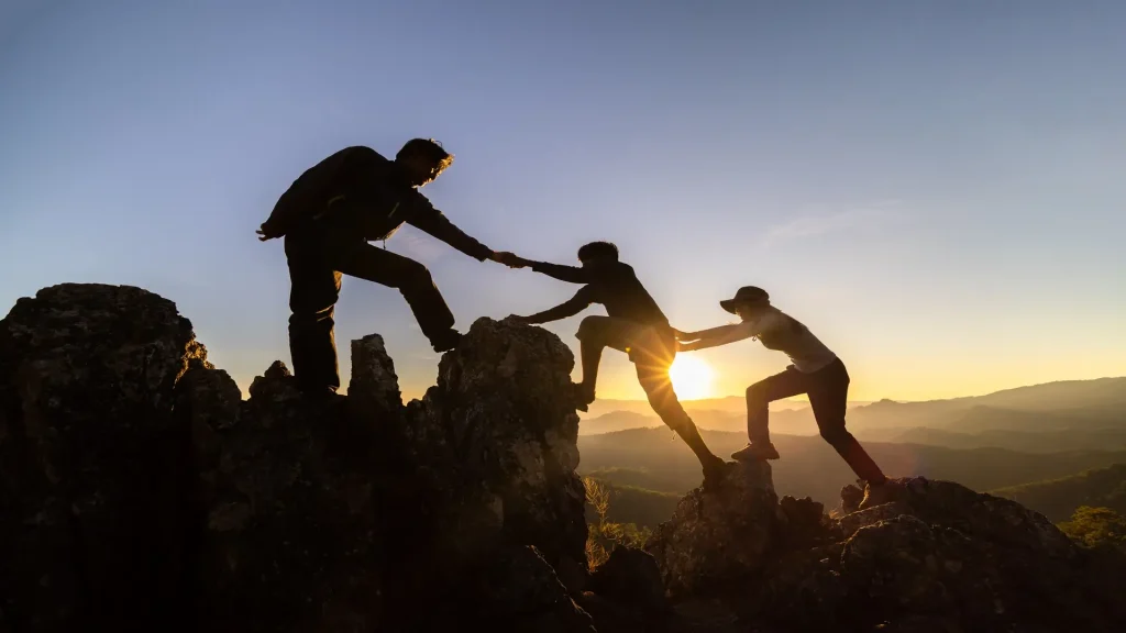 Three hikers silhouetted at sunrise, helping each other in a trust climb on a rugged mountain peak.