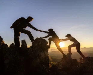 Three hikers silhouetted at sunrise, helping each other in a trust climb on a rugged mountain peak.