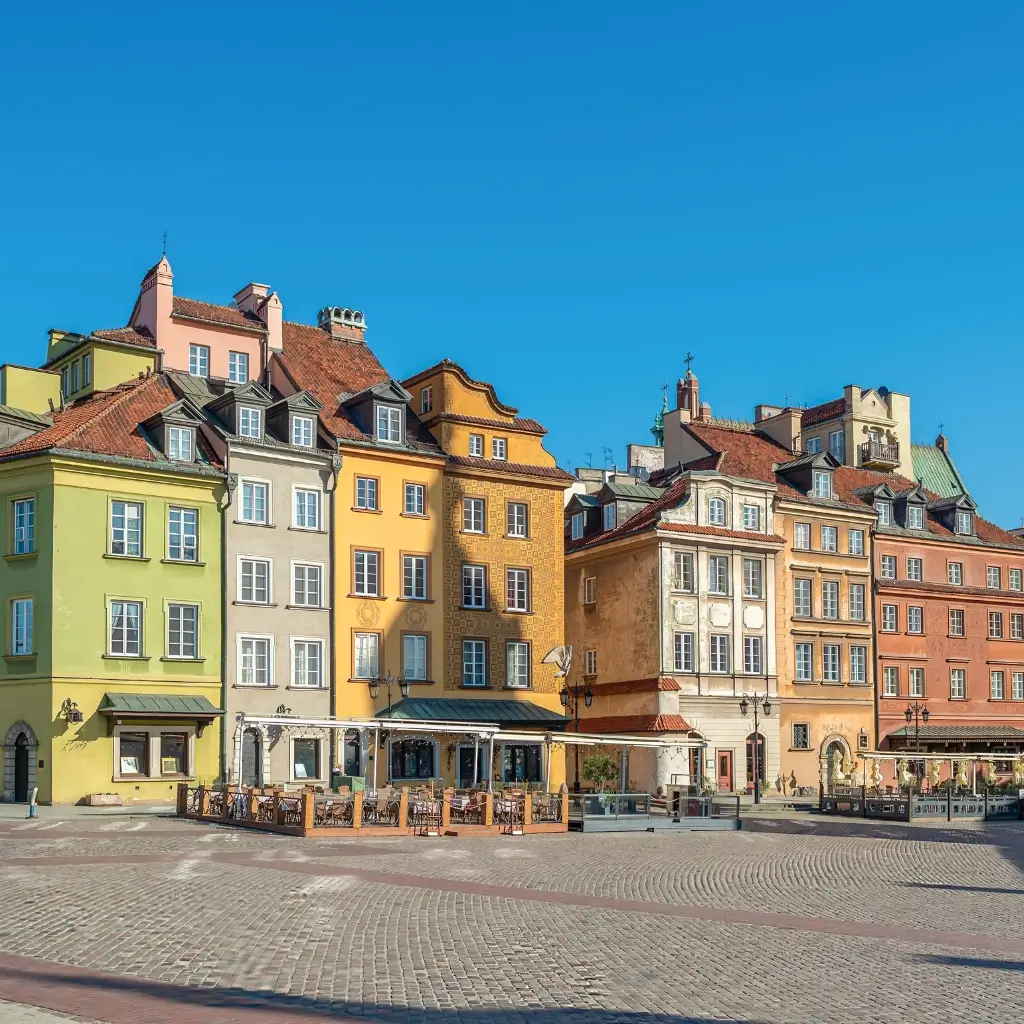 A wide cobblestone square lined with historic, brightly colored buildings under a clear blue sky in Warsaw, Poland.