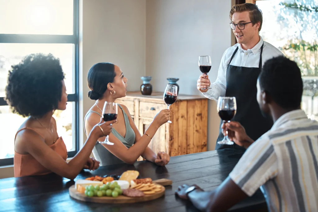 Professional sommelier presenting red wine to a group of friends seated around a table with a cheese board.