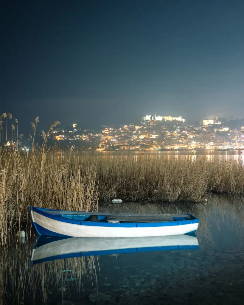 A quiet blue rowboat resting on the tranquil waters of Lake Ohrid at night, with an illuminated historic European town and fortress in the background, representing the serene nature and history found on our custom cruise and tour itineraries.